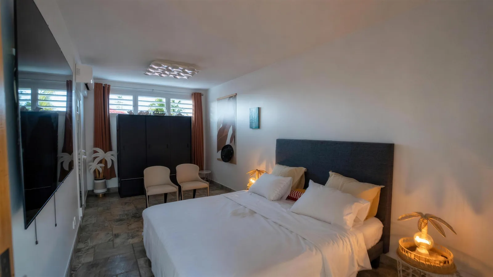 Chambre Palms Beach — lit, lampes palmier, fauteuils crème et grand écran, villa House of Harmony, Goyave